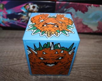 May include: A blue cube with a monster design on each side. The top side features a green and orange monster with its mouth open. The bottom side features a green and orange monster with its mouth closed.