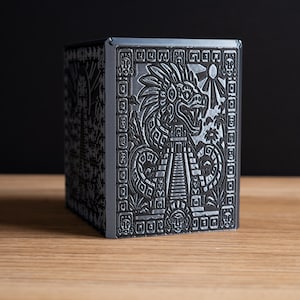 May include: A dark gray, rectangular, box-shaped object featuring intricate, raised relief carvings on its side. The design depicts a feathered serpent deity, a stepped pyramid, and sun rays, all framed by a decorative border of geometric symbols.