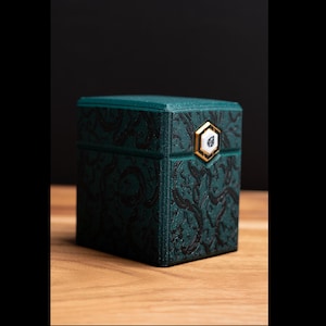 May include: A teal-colored box with a textured, swirling pattern. The box has a gold-colored hexagonal clasp with a white center. The box is sitting on a wooden surface.