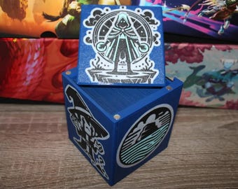 May include: A blue wooden box with a hinged lid. The lid has a white and blue graphic of a person in a hooded cloak. The box has a white and blue graphic of a person in a hat and a white and blue graphic of a mountain range.