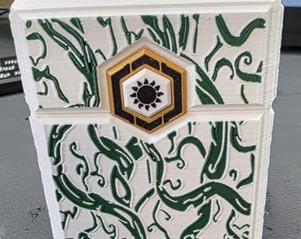 May include: A white box with green vine-like patterns and a gold and black hexagonal emblem. The emblem features a sun-like design in the center. The box has a textured surface and appears to be 3D-printed.