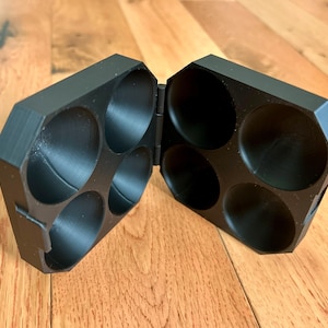 May include: A black, hinged container with eight circular compartments. The container is open, revealing the interior. The exterior has a geometric, faceted design. The object is made of a matte black material and sits on a wooden surface.