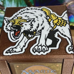 May include: A brown card storage box featuring a white wolf illustration with gold accents. The wolf is snarling, with its mouth open, revealing a red tongue. The box is designed for collectible cards.