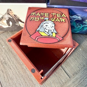 May include: A red square box with a hinged lid, featuring the text "MAKE TEA NOT WAR" and an illustration of a character. The box is open, revealing a compartment and a silver tool. Other boxes with fantasy art are in the background.
