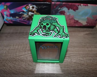 May include: Green deck box with a creature illustration on the lid. The creature is a green, tree-like figure with a face and arms. The box has a clear window on the front, showing a "Magic: The Gathering" card.