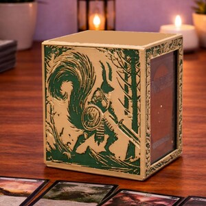 May include: A beige card storage box with a green illustration of a squirrel warrior in a forest scene. The box is rectangular and has a hinged lid. Several trading cards are visible in the foreground.