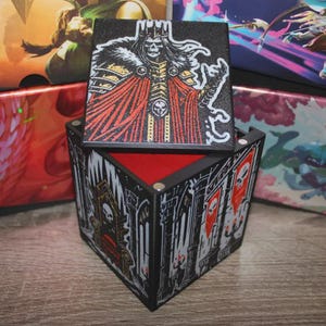 May include: A black, angular dice box with a red interior and a removable lid. The lid features a skeletal king illustration in red, gold, and black. The box exterior displays a gothic castle design with skull banners.