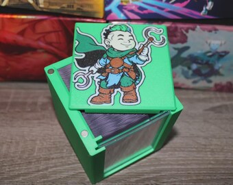 May include: A green card storage box with a hinged lid. The lid features a cartoon illustration of a character with a staff. The box is open, revealing the interior and a stack of cards. The box is made of plastic.