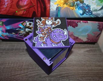 May include: A purple card holder with a hinged lid, holding a stack of cards. The lid features a cartoon illustration of a character riding a purple rhinoceros. The background shows other card holders with colorful designs.