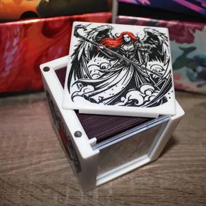 May include: White cube-shaped box with a clear lid. The lid features a black and white illustration of a woman with red hair and wings. The woman is holding a staff. The box is sitting on a wooden surface.