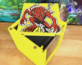 May include: A yellow, geometric card box with a red and black cartoon demon graphic on the lid. The box is open, revealing a stack of black cards inside. The box is made of wood.