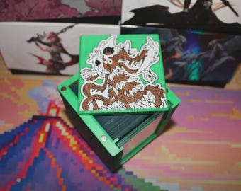 May include: Green and black card storage box with a green lid featuring a brown and white monster illustration. The box is open, revealing a stack of cards. Other boxes with illustrations are in the background.