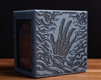 May include: A gray, cube-shaped card storage box with a detailed relief carving of a skeletal hand reaching out from a swirling, organic background. The box has a card visible on one side.