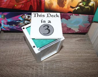 May include: A white card holder with a stack of green cards inside. A white card on top reads "This Deck is a 3" in black text. The holder is on a wooden surface, with other boxes in the background.