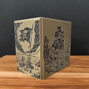 May include: A gold-colored, cube-shaped box with detailed black engravings. One side depicts a pirate ship sailing on the sea, while another shows a pirate flag with a skull and crossbones. The box is set on a wooden surface.