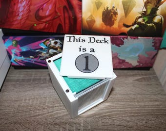 May include: A white card with the text "This Deck is a 1" sits atop a white box filled with teal cards. The box is on a wooden surface, with colorful boxes in the background. The image is of a card game.