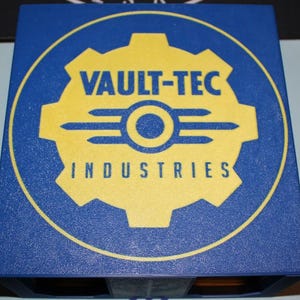 May include: A blue box with a yellow Vault-Tec Industries logo. The logo features a gear shape with the text "VAULT-TEC" above a stylized symbol and the word "INDUSTRIES" below. The box has a textured surface.