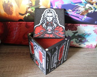May include: A black wooden box with a red interior and a removable lid. The lid features a colorful illustration of a man with long black hair and a red cape. The box has a gothic design with black and red accents.