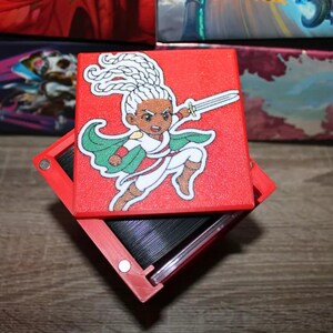 May include: A red box with a hinged lid. The lid features a colorful illustration of a cartoon character with white hair, a green cape, and a sword. The character is wearing a white shirt and pants.