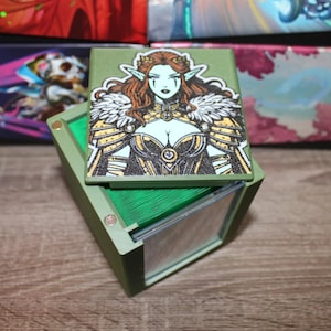 May include: A green card deck box with a removable lid featuring a colorful illustration of a woman with green skin and long blonde hair wearing a gold and white armor. The box is open and shows the green card deck inside.