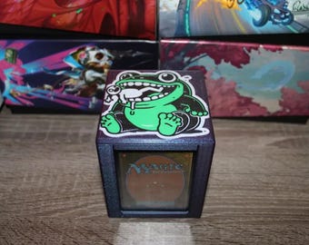 May include: A dark blue card storage box with a green frog sticker on top. The frog has a white creature in its mouth. The box has a card visible through a window. Other boxes with colorful designs are in the background.