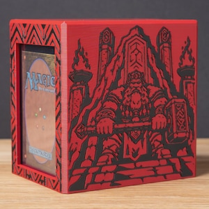 May include: A red and black rectangular deck box featuring a 3D-printed relief of a bearded figure sitting on a throne holding a large hammer. The front frame displays a Magic The Gathering card with a visible back design and the text Deckmaster.