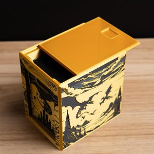 May include: A rectangular yellow storage box with a sliding lid, featuring black relief carvings on the sides that depict silhouettes of flying dragons, jagged mountain peaks, and tall castle towers against a cloudy sky background.