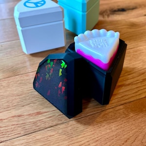 May include: A black geometric box with a holographic red and green inlay, containing a white and pink plastic cake slice with the text "NIPP DOH" embossed on top. Two additional boxes, one white with a blue peace sign and one light blue, sit behind.