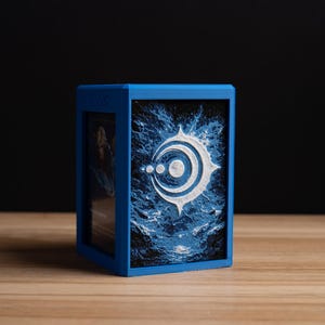 May include: A blue card storage box with a white and blue graphic design. The box is rectangular and sits on a wooden surface. The graphic features a stylized emblem with a circular design.