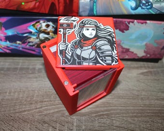May include: Red card deck box with a removable lid featuring a black and white illustration of a woman with a red scarf holding a hammer. The box is made of plastic and has a clear window on the front.