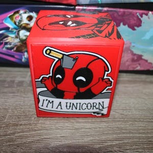 May include: A red cube-shaped box with a cartoon image of Deadpool with a unicorn horn and the text "I'm a unicorn".