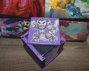 May include: A purple card storage box with a matching lid featuring a cartoon warrior illustration. The box is open, revealing a stack of cards inside. The background includes other boxes with colorful artwork.