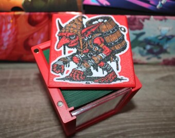 May include: Red card storage box with a hinged lid. The lid features a sticker of a red goblin carrying a barrel. The box is open, revealing green cards inside. The box is designed for trading cards.
