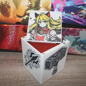 May include: A white, cube-shaped box with a red interior, featuring illustrations of a female anime character and a hammer. The box is decorated with black and white graphics of a hammer and a weapon. The box is sitting on a wooden surface.