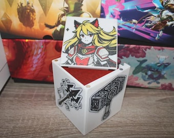 May include: A white, cube-shaped box with a red interior, featuring illustrations of a female anime character and a hammer. The box is decorated with black and white graphics of a hammer and a weapon. The box is sitting on a wooden surface.