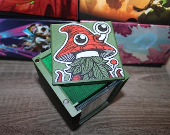 May include: Green card deck box with a removable lid featuring a cartoon illustration of a mushroom creature with a red cap, white eyes, and a green beard. The box is made of sturdy plastic and has a clear window on the front.