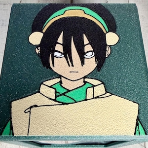 May include: A green box featuring a cartoon illustration of a character with black hair, white eyes, and a green headband. The character is wearing a tan jacket over a green shirt. The box has a textured surface.