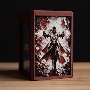 May include: A burgundy card storage box featuring a League of Legends design. The front panel displays a detailed illustration of a character with glowing hands. The box is made of wood and sits on a wooden surface.