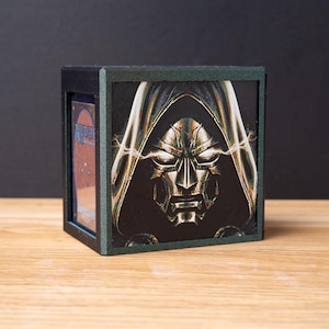 May include: A square black card storage box featuring a graphic illustration of Doctor Doom on the front panel. The character wears a metallic mask and a dark hood with glowing yellow energy effects. A Magic The Gathering card is visible inside.