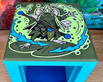 May include: A square, light blue table with a detailed illustration on the top. The illustration features a green frog with a staff, surrounded by green and blue water-like elements. The frog is on a lily pad.