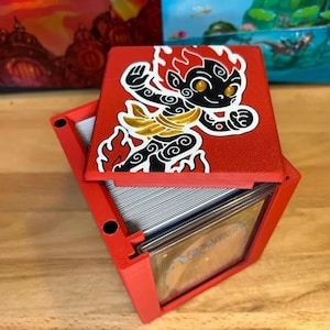 May include: Red card storage box with a hinged lid. The lid features a cartoon character design in black, gold, and white. The box is open, revealing a stack of cards inside. The box is sitting on a wooden surface.