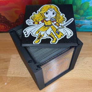 May include: Black card storage box with a removable lid featuring a cartoon illustration of a female warrior with blonde hair, a sword, and yellow and white clothing. The box is open, revealing a stack of cards.