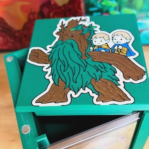 May include: A green card storage box with a hinged lid. The lid features a sticker of a cartoonish, tree-like creature with two small figures riding on its back. The box is open, revealing the interior.
