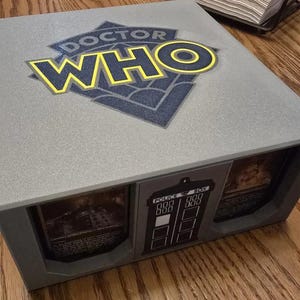May include: A gray box with a Doctor Who logo on the top. The box has two compartments with images of the TARDIS and a Dalek inside. The box is made of plastic and has a textured surface.