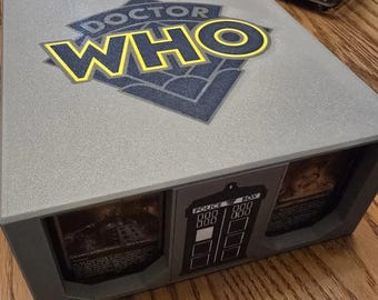 May include: A gray box with a Doctor Who logo on the top. The box has two compartments with images of the TARDIS and a Dalek inside. The box is made of plastic and has a textured surface.