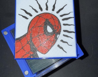 May include: A blue and white case featuring a red and black illustration of Spider-Man's head. The artwork is on a white background with black sunburst lines. The case is open, revealing a clear interior.