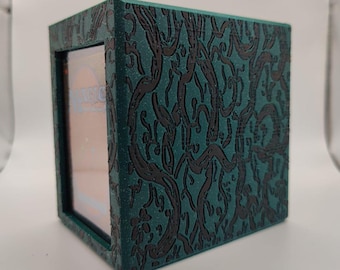 May include: A teal and black card storage box with a decorative, swirling pattern. The box has a rectangular window on one side, and the word "MAGIC" is visible through the window. The box is designed to hold cards.