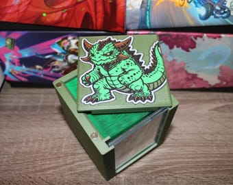 May include: A green card storage box with a hinged lid, featuring a cartoon dragon sticker. The box is olive green with a clear window. The dragon sticker is green with brown accents. The box is designed to hold cards.