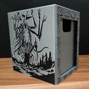 May include: A gray, cube-shaped storage box with a detailed black illustration of a creature and a cityscape. The box has a handle cutout and decorative border. The design is 3D printed.