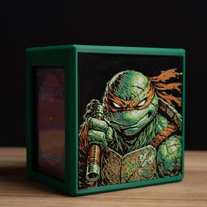 May include: Green card storage box featuring a Teenage Mutant Ninja Turtle illustration. The box has a green frame and a black background for the image. The turtle is holding a weapon and has an orange mask.